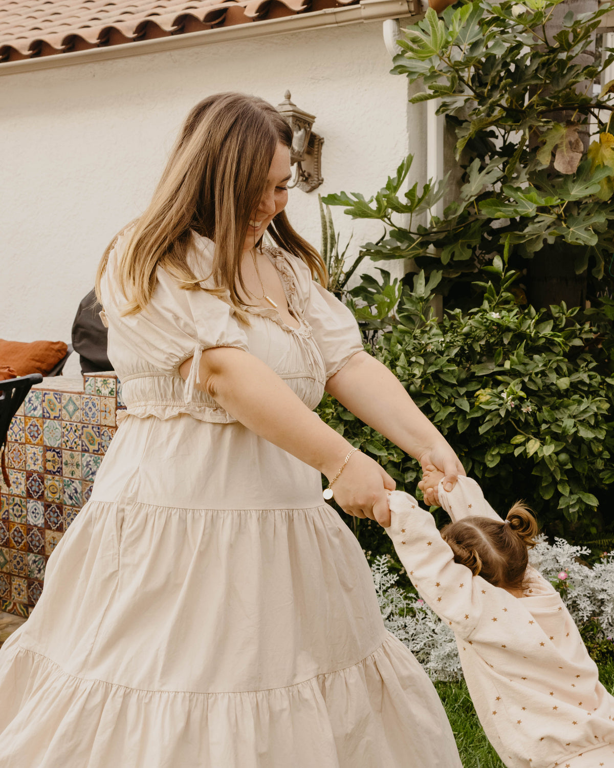 A woman in a cream-colored tiered puff-sleeve dress helps a young child wearing a matching cream polka-dot top in an outdoor garden setting.