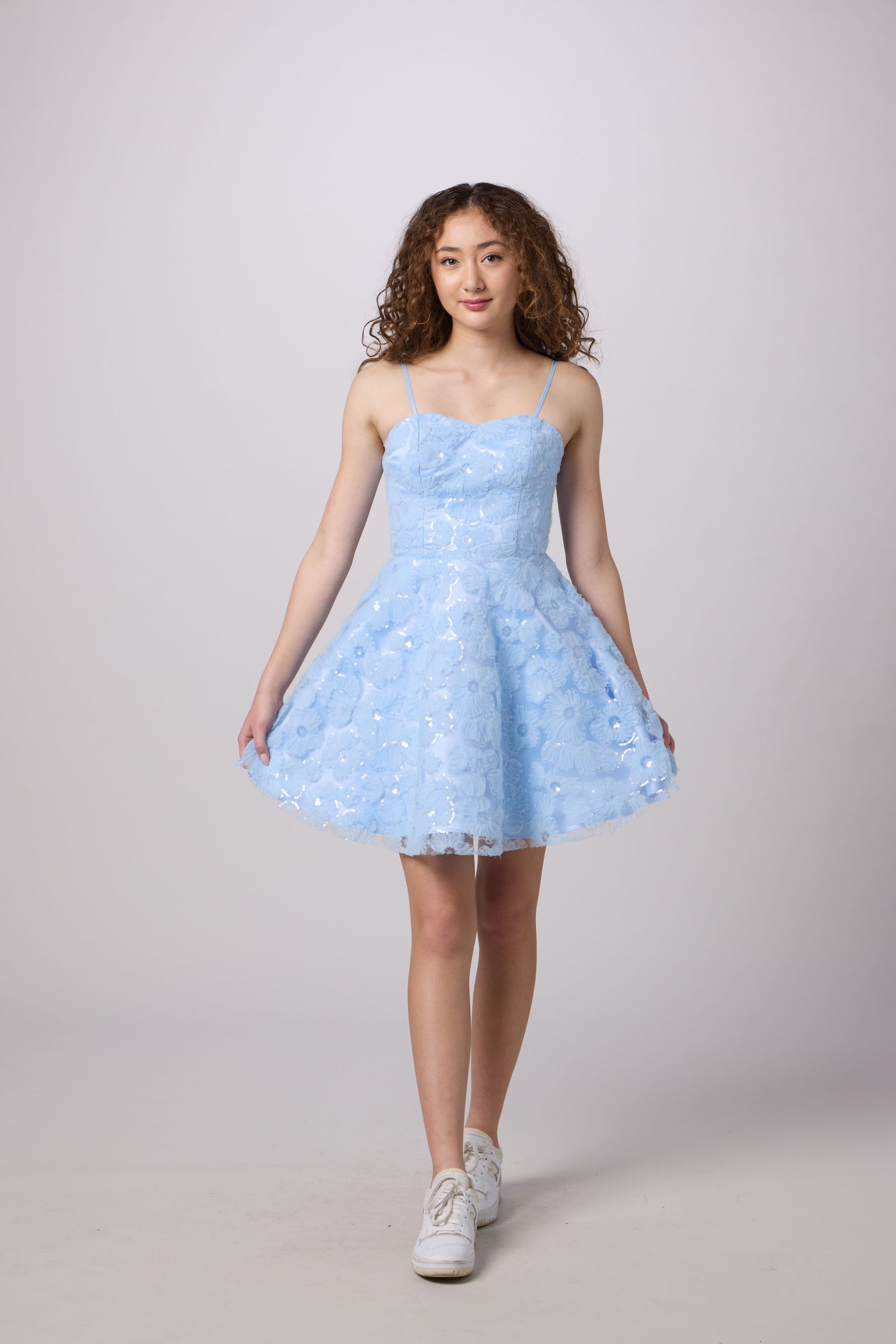 A young woman modeling a light blue sleeveless A-line dress with a textured floral lace bodice and a flared tulle skirt, paired with white sneakers, against a solid light gray studio background.