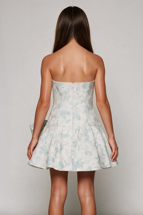 A woman shown from the back wearing a strapless mini dress with a light blue and white floral pattern, photographed against a neutral light gray studio background.