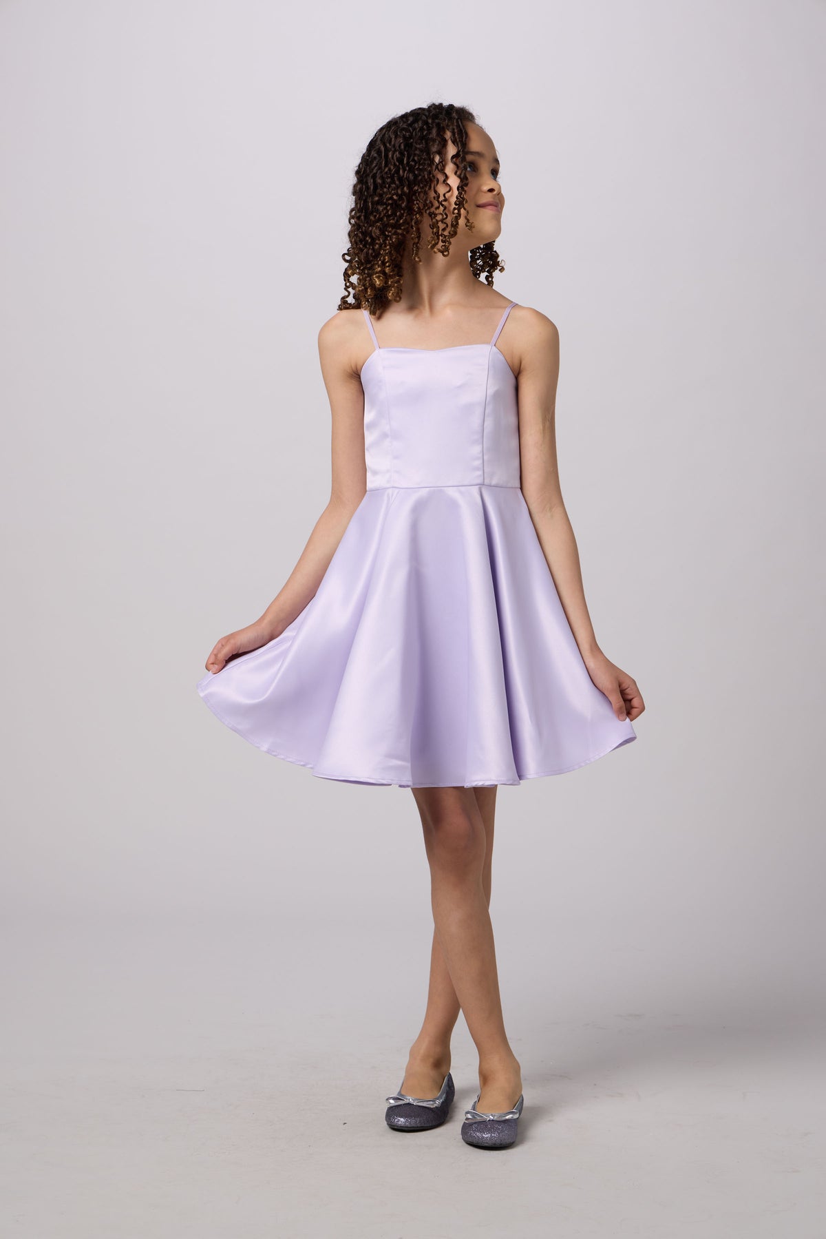 A young girl with curly brown hair wearing a light lavender satin spaghetti-strap dress and silver glitter flats, standing against a light gray studio background.