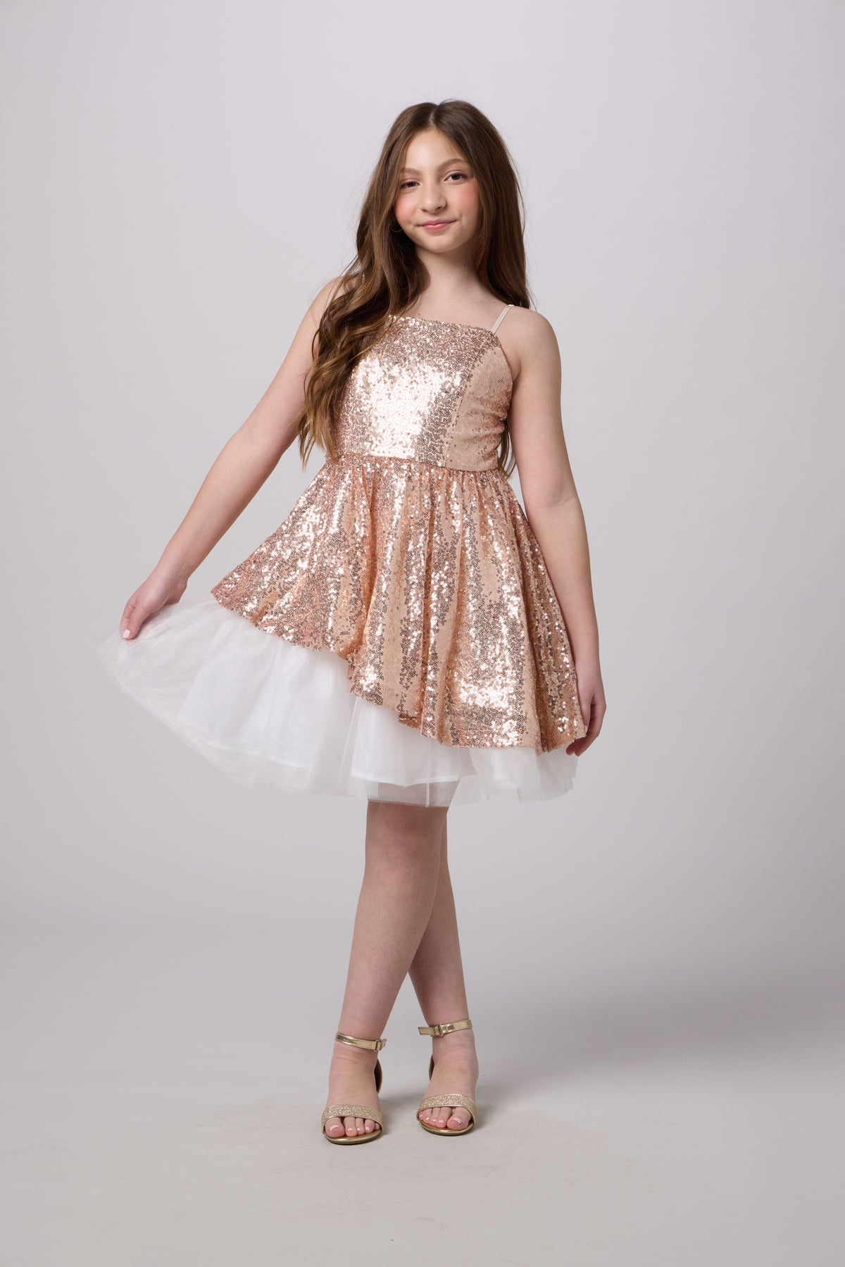 A young girl models a strapless sequined dress with a rose gold sequin bodice and a layered white tulle skirt, paired with gold sandals, against a light gray studio background.