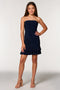 A young woman modeling a strapless navy blue mini dress with ruching and a ruffled hem, paired with beige high-heeled sandals, against a light gray studio background.