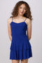 A woman modeling a bright royal blue spaghetti-strap mini dress with a tiered ruffle skirt, paired with silver heeled sandals, against a light gray studio background.