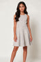 A sleeveless fit-and-flare dress with a small white grid pattern on a light beige background, modeled by a young woman against a plain light gray studio backdrop.