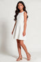 A young girl with long dark hair models a sleeveless white textured mini dress with a flared skirt, paired with beige ankle-strap block-heel sandals, against a plain light gray studio background.
