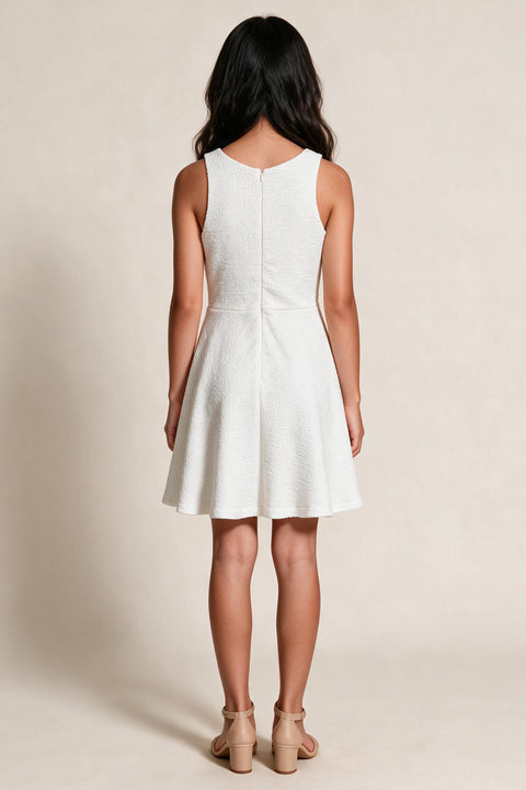 A young girl viewed from the back wearing a sleeveless white fit-and-flare dress with a textured fabric and a V-back design, paired with beige high-heeled sandals against a warm beige studio background.