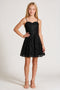 A young girl with blonde hair wearing a black spaghetti-strap mini dress with sequins and a flared skirt, paired with nude high-heeled sandals, standing against a light gray studio background.