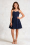A young girl modeling a navy blue lace spaghetti-strap mini dress with a sweetheart neckline and flared skirt, paired with beige strappy high-heeled sandals, against a textured light gray studio backdrop.