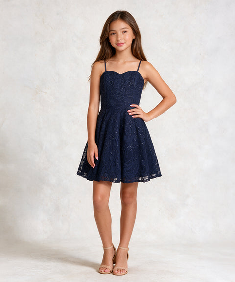 A young girl modeling a navy blue lace spaghetti-strap mini dress with a sweetheart neckline and flared skirt, paired with beige strappy high-heeled sandals, against a textured light gray studio backdrop.