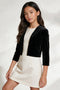 A young girl modeling a black velvet cropped jacket over a cream-colored dress, photographed against a light studio background with soft shadows.