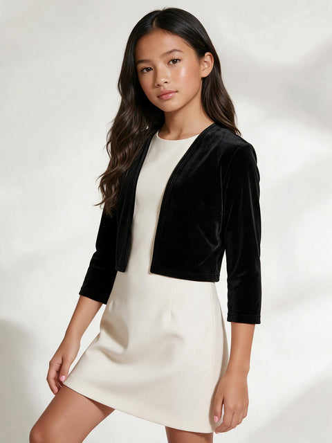 A young girl modeling a black velvet cropped jacket over a cream-colored dress, photographed against a light studio background with soft shadows.
