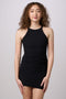 A woman with curly brown hair wearing a black ribbed halter-neck mini dress, standing against a solid light gray studio background.