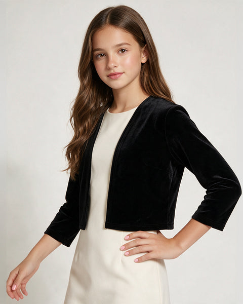 A young girl with long brown hair models a black velvet cropped jacket over a cream-colored dress against a light gray studio background.