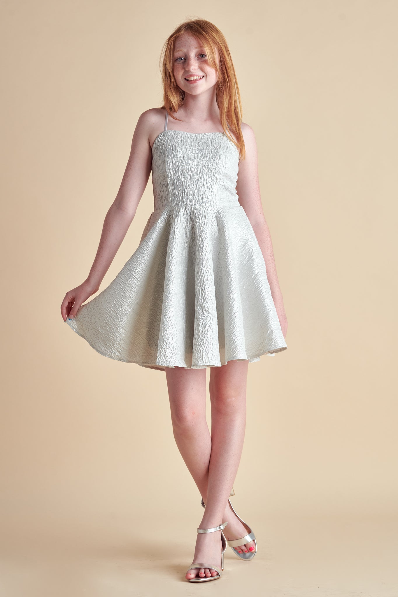 A red haired girl in an all over non-stretch, texture fabric dress made in a longer length in silver. It hits right above the knee and features a full circle skirt.