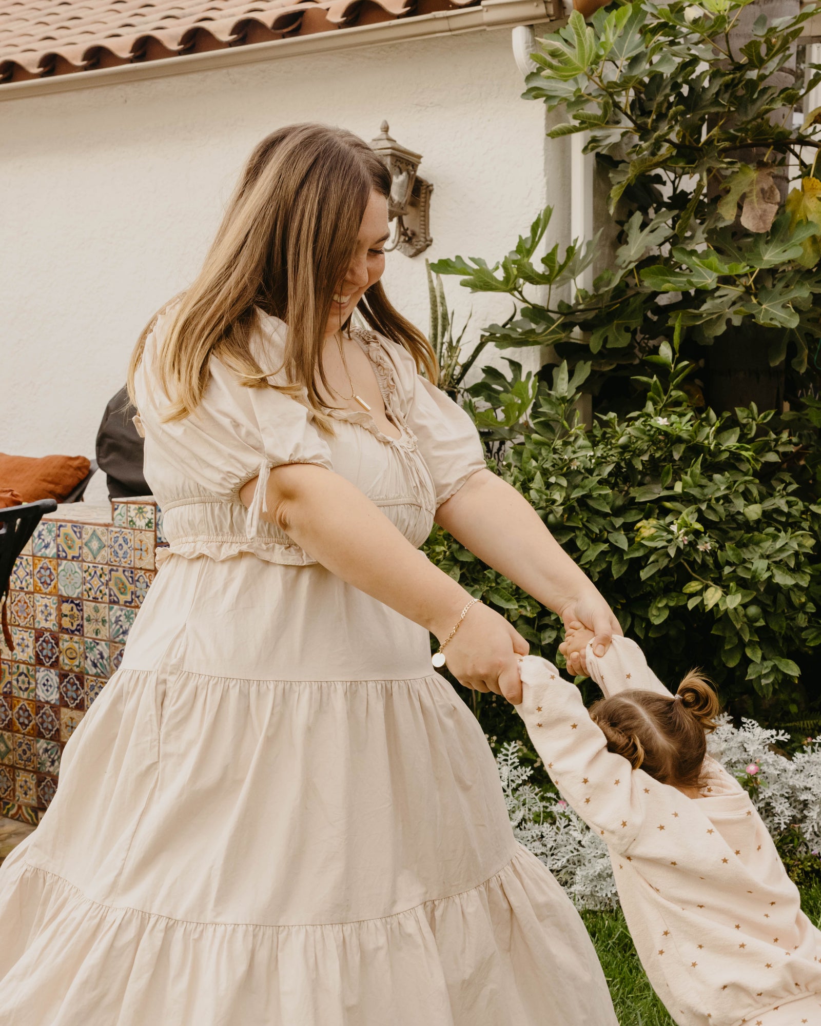 A woman in a cream-colored tiered puff-sleeve dress helps a young child wearing a matching cream polka-dot top in an outdoor garden setting.