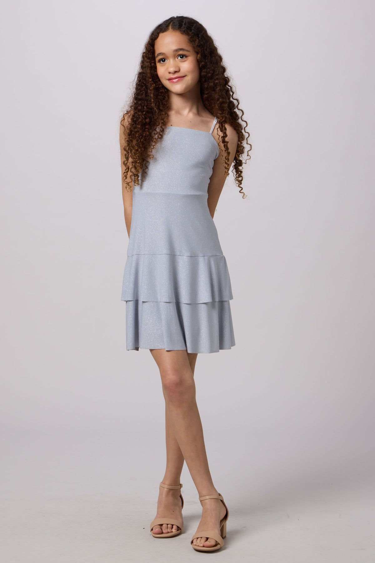 A young girl with long, curly brown hair stands against a light gray studio background wearing a light blue sleeveless tiered mini dress and beige heeled sandals.