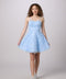 A young woman modeling a light blue sleeveless A-line dress with a textured floral lace bodice and a flared tulle skirt, paired with white sneakers, against a solid light gray studio background.