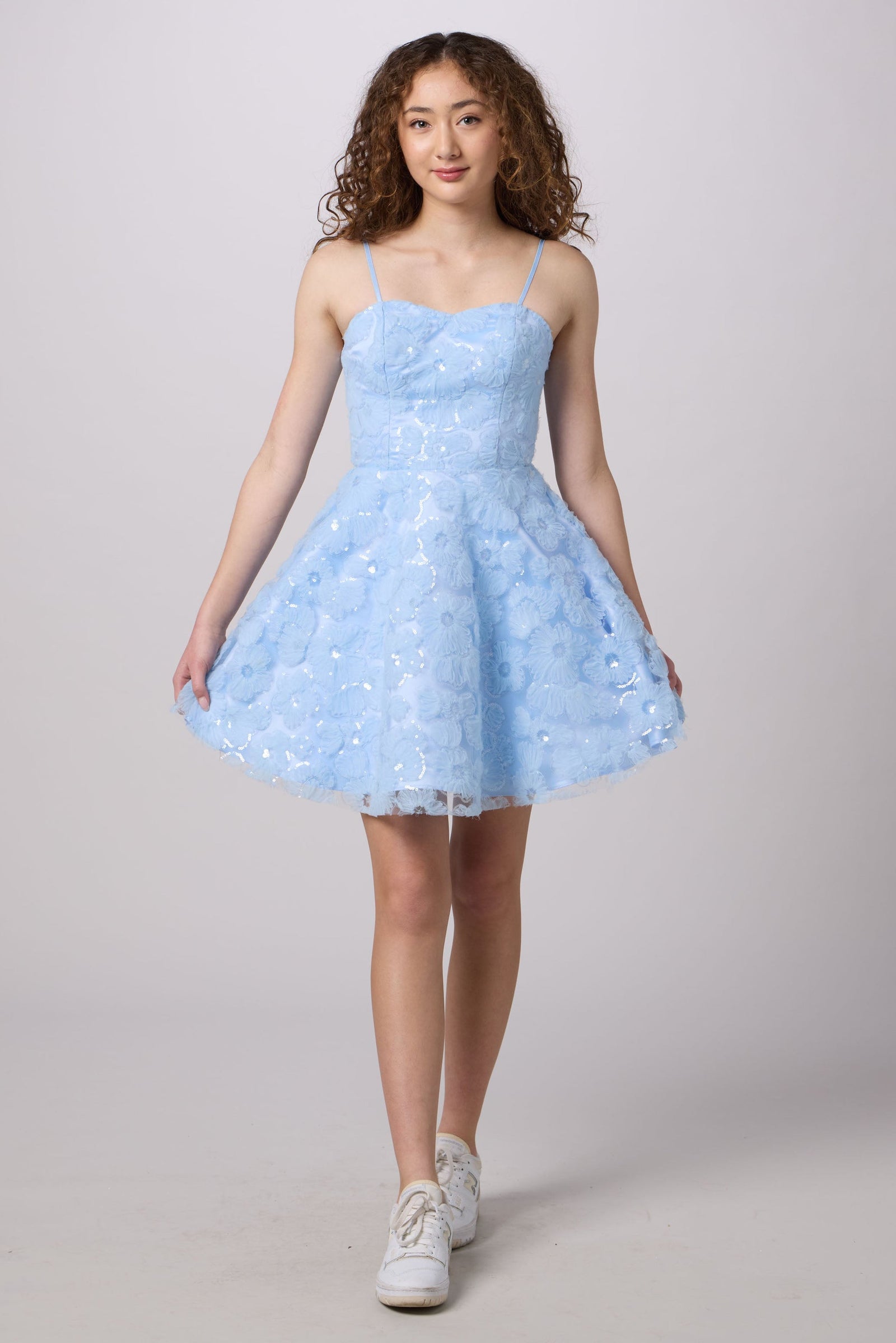 A young woman modeling a light blue sleeveless A-line dress with a textured floral lace bodice and a flared tulle skirt, paired with white sneakers, against a solid light gray studio background.