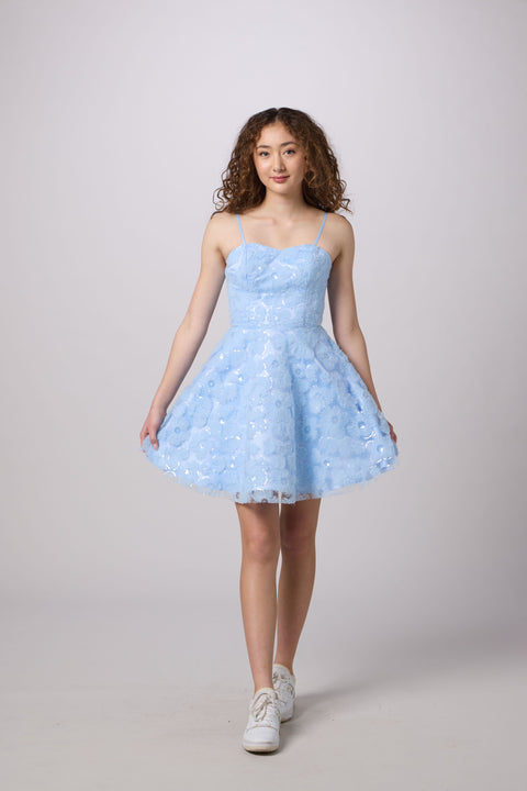 A young woman modeling a light blue sleeveless A-line dress with a textured floral lace bodice and a flared tulle skirt, paired with white sneakers, against a solid light gray studio background.