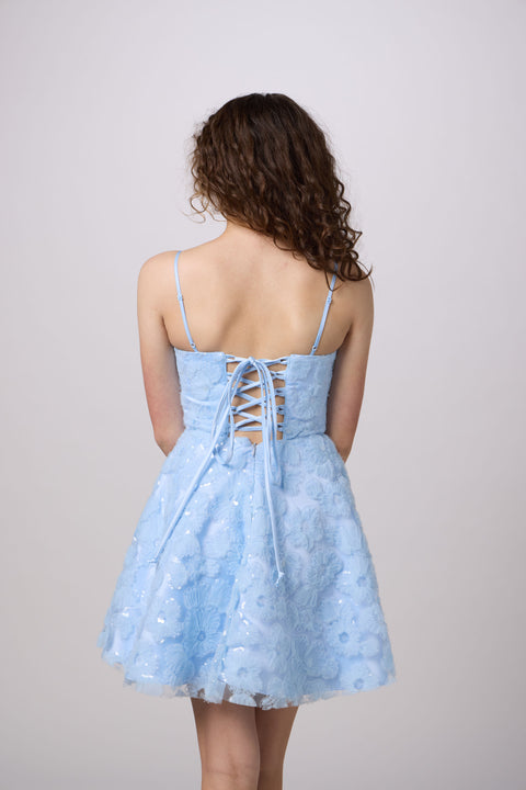 A young woman viewed from behind wearing a light blue lace-up mini dress with spaghetti straps and a flared tulle skirt, photographed against a plain light gray studio background.