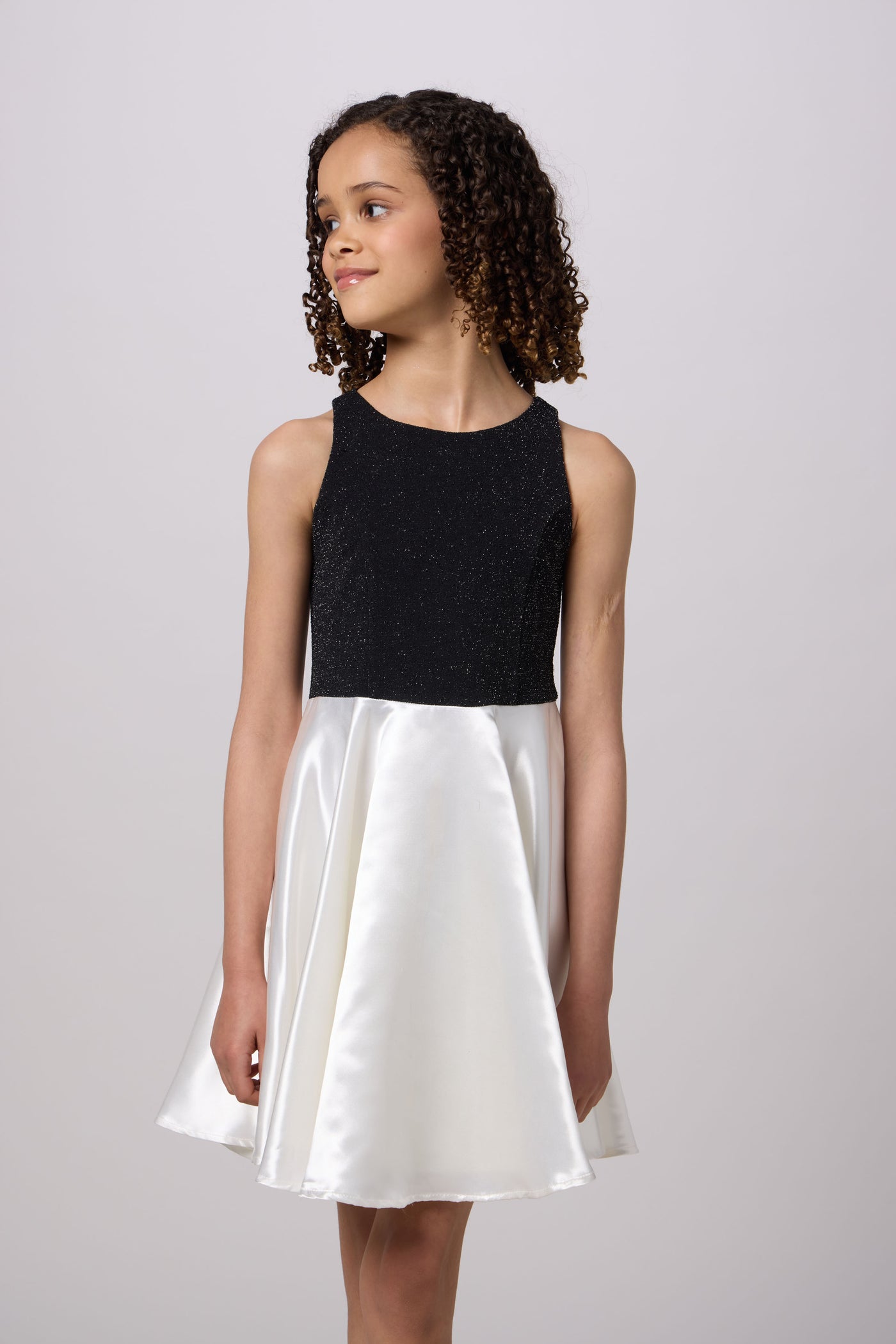 A young girl with curly brown hair wearing a sleeveless two-tone dress with a black textured bodice and a white satin skirt, standing against a light gray studio background.