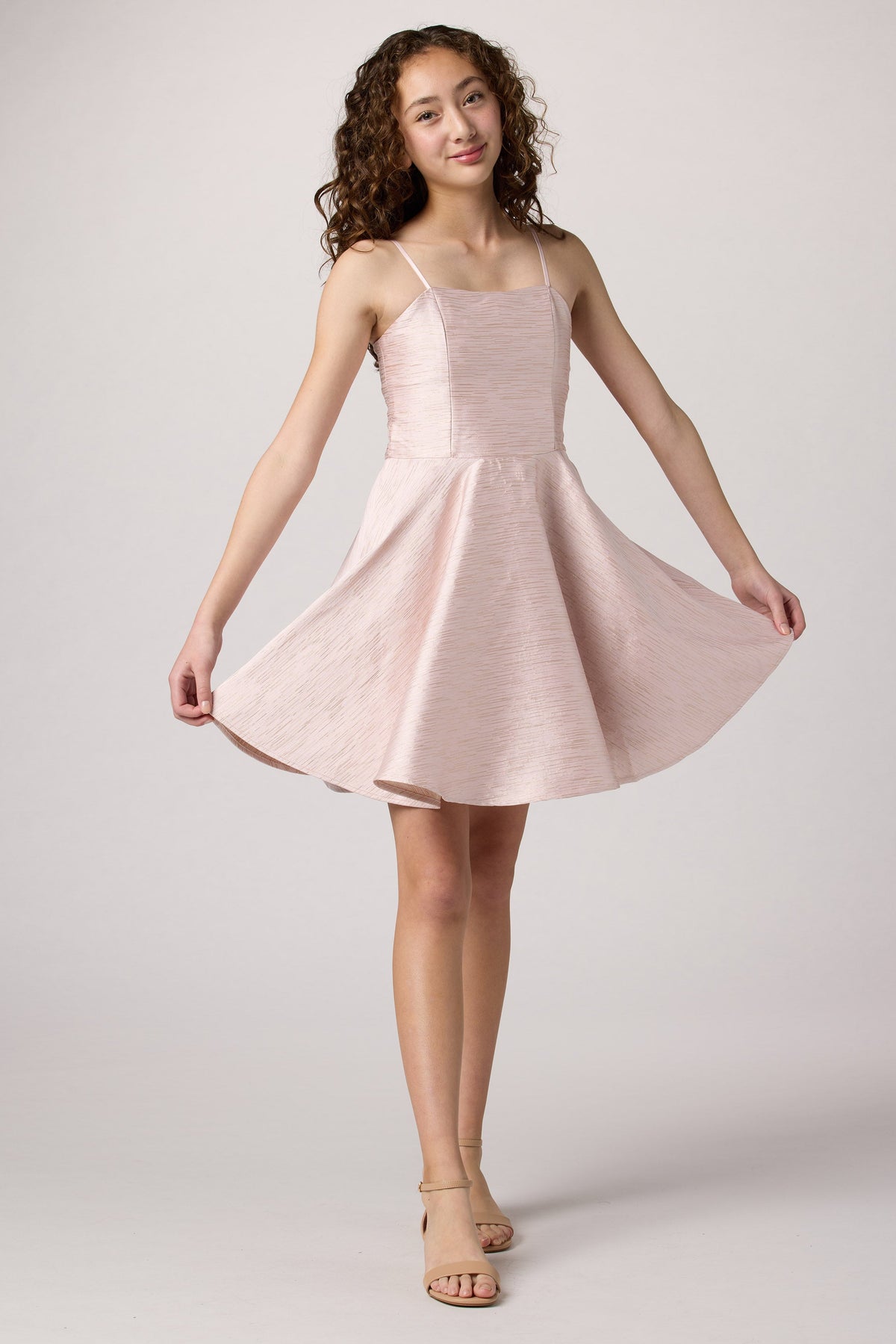 A young woman with curly brown hair models a light pink spaghetti-strap mini dress with a flared skirt, holding out the sides of the skirt to show its volume against a neutral light gray studio background.