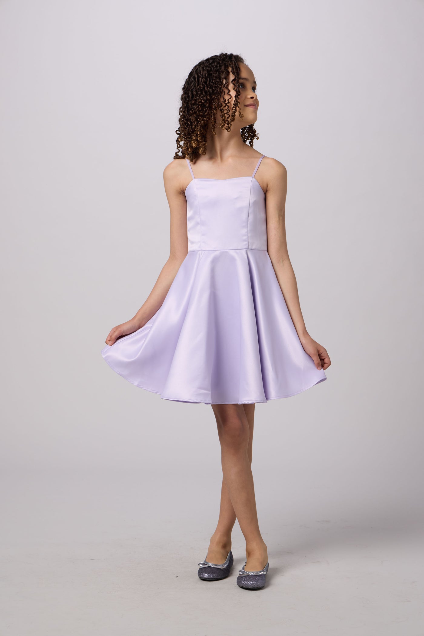 A young girl with curly brown hair wearing a light lavender satin spaghetti-strap dress and silver glitter flats, standing against a light gray studio background.