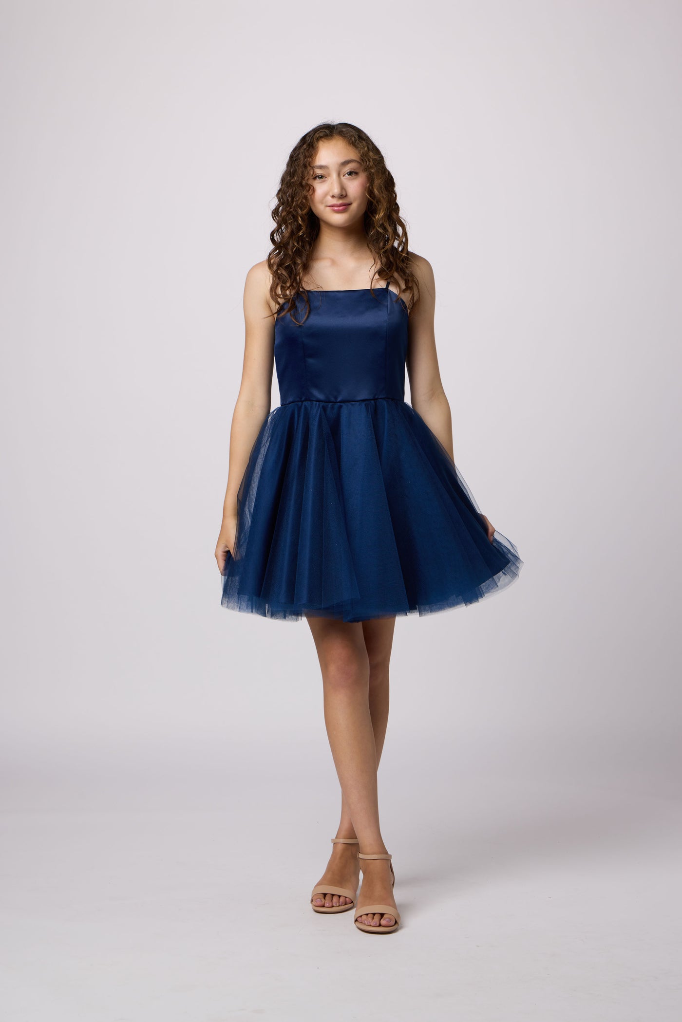 A young woman modeling a short navy blue tulle dress with a fitted bodice and a flared skirt, standing against a light gray studio background.