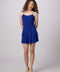 A woman modeling a bright royal blue spaghetti-strap mini dress with a tiered ruffle skirt, paired with silver heeled sandals, against a light gray studio background.