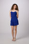 A woman modeling a bright royal blue spaghetti-strap mini dress with a tiered ruffle skirt, paired with silver heeled sandals, against a light gray studio background.