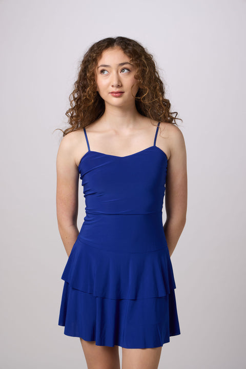 A young woman with curly brown hair wearing a royal blue tiered spaghetti-strap dress, standing against a light gray studio background.