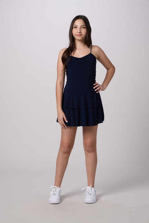 A young woman modeling a navy blue sleeveless mini dress with a tiered skirt, paired with white sneakers, against a light gray studio background.