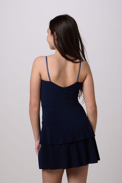 A woman shown from the back wearing a navy blue sleeveless mini dress with thin straps and a ruffled tiered skirt, photographed against a plain light gray studio background.