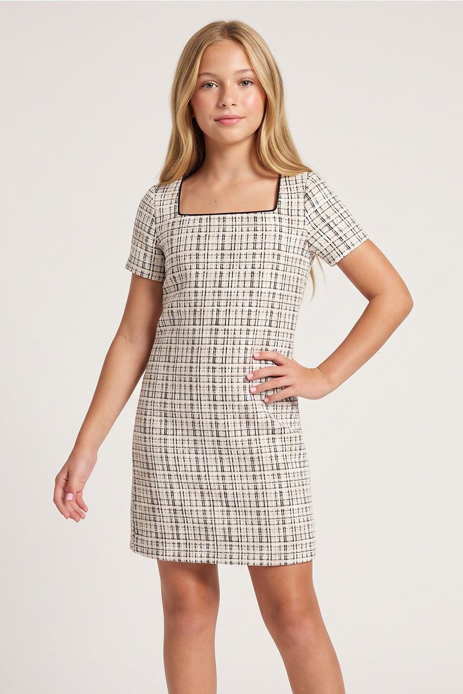 A young woman modeling a short-sleeve, knee-length A-line dress with a beige and black windowpane plaid pattern, set against a plain light gray studio background.