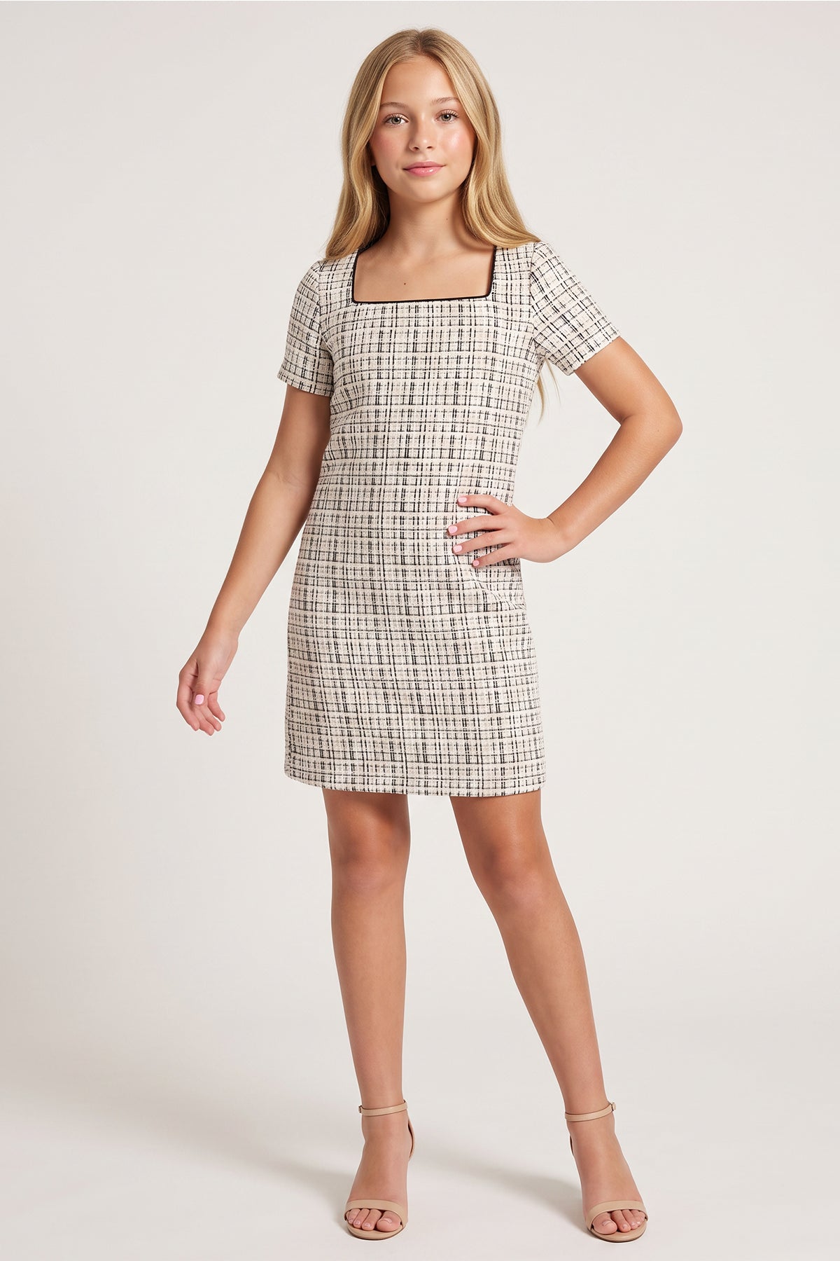 A young woman modeling a short-sleeve, knee-length A-line dress with a beige and black windowpane plaid pattern, set against a plain light gray studio background.