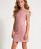 A young girl with long wavy blonde hair wearing a textured dusty pink sleeveless fit-and-flare dress, standing against a light gray textured studio background.