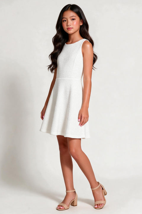 A young girl with long dark hair models a sleeveless white textured mini dress with a flared skirt, paired with beige ankle-strap block-heel sandals, against a plain light gray studio background.