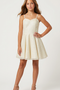 A young girl modeling a short, sleeveless cream dress with thin spaghetti straps, a sweetheart neckline, and a flared skirt, paired with nude high-heeled sandals against a light gray studio background.