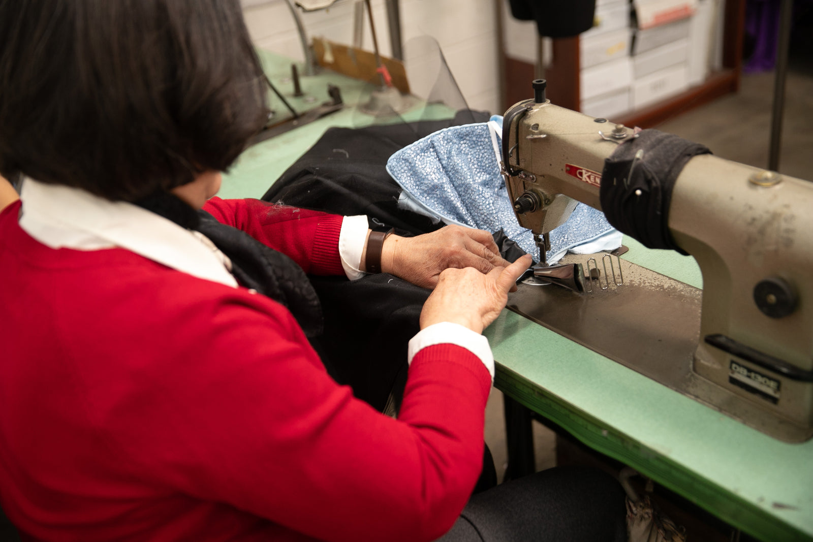 A person in a red sweater sits at a sewing machine, stitching a black garment with a light blue fabric piece. The scene is set in a workshop with sewing equipment and materials on a green table.