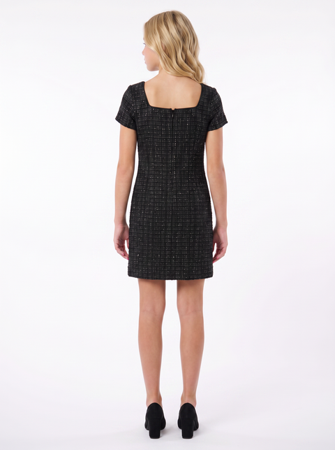 A woman viewed from the back wearing a short-sleeve black tweed dress with a square neckline and black heels, photographed against a plain white studio background.