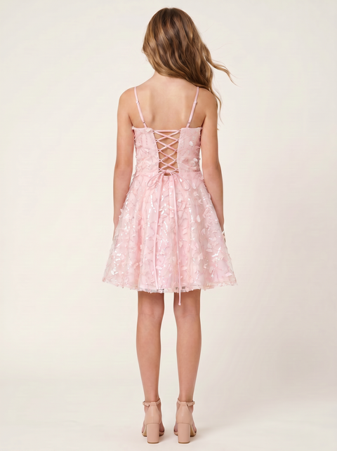 A young woman modeling a light pink sleeveless fit-and-flare dress with thin straps and a flared skirt, paired with nude high-heeled sandals, against a plain light beige studio background.