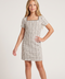 A young girl models a short-sleeve, knee-length shift dress with a beige and black plaid pattern and a square neckline, paired with beige strappy heels, against a plain light gray studio background.