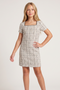 A young girl models a short-sleeve, knee-length shift dress with a beige and black plaid pattern and a square neckline, paired with beige strappy heels, against a plain light gray studio background.