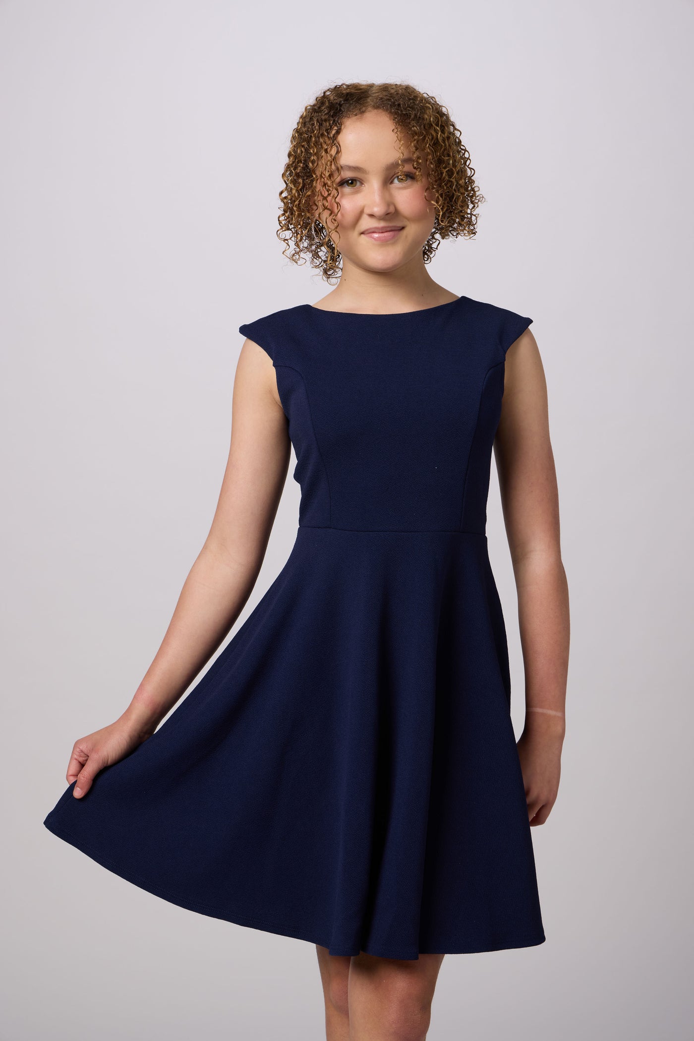 A young girl with curly brown hair wearing a sleeveless navy blue A-line dress, standing against a light gray studio background.