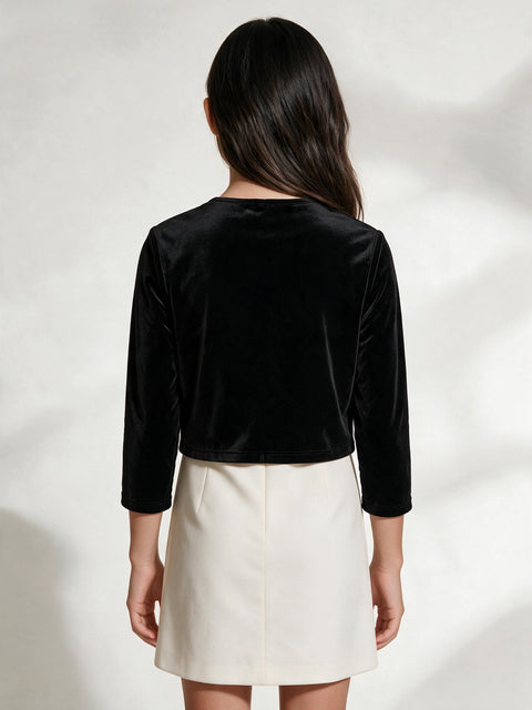 A model shown from the back wearing a black velvet long-sleeve cropped top and a cream-colored mini skirt, set against a light gray background with soft leaf shadows.
