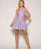 A young girl with long blonde hair models a strapless lavender and white patterned mini dress with a flared skirt, paired with beige high-heeled sandals, against a plain light gray studio background.