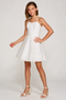 A young girl modeling a white spaghetti-strap mini dress with a flared skirt, paired with beige block-heel sandals, against a light gray studio background.