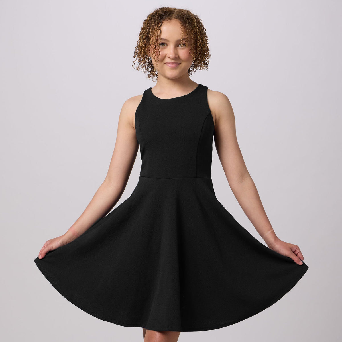 A young girl with curly brown hair wearing a sleeveless black skater dress and silver heeled sandals, posing in a studio setting against a light gray background.