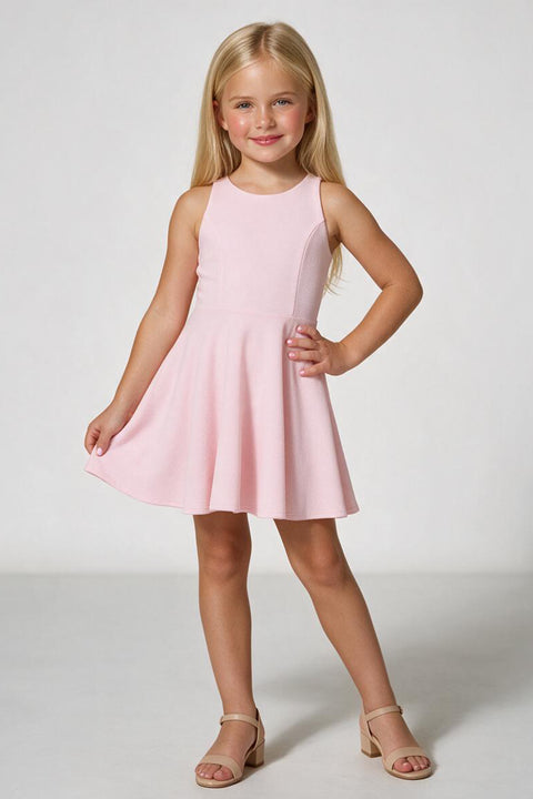A young girl with blonde hair stands in a studio wearing a sleeveless light pink dress with a flared skirt and beige heeled sandals against a light gray background.