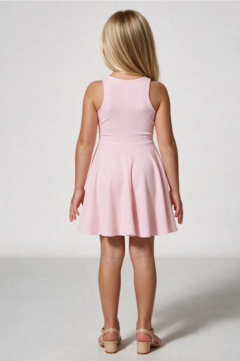 A young girl with blonde hair is shown from the back wearing a sleeveless light pink skater dress and beige sandals, standing against a light gray studio background.